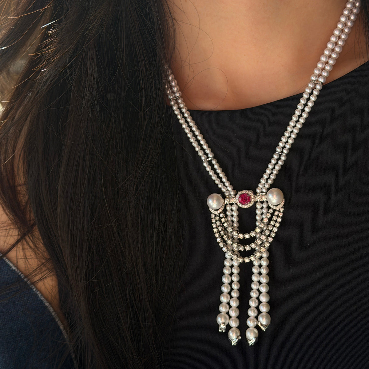 A model wearing a luxurious double-strand Akoya pearl necklace featuring a versatile centerpiece brooch. The intricate brooch in IMG_0810.jpg showcases a 1.16ct ruby surrounded by a diamond halo, flanked by two large pearls, with cascading diamond swags and three Akoya pearl tassel drops.