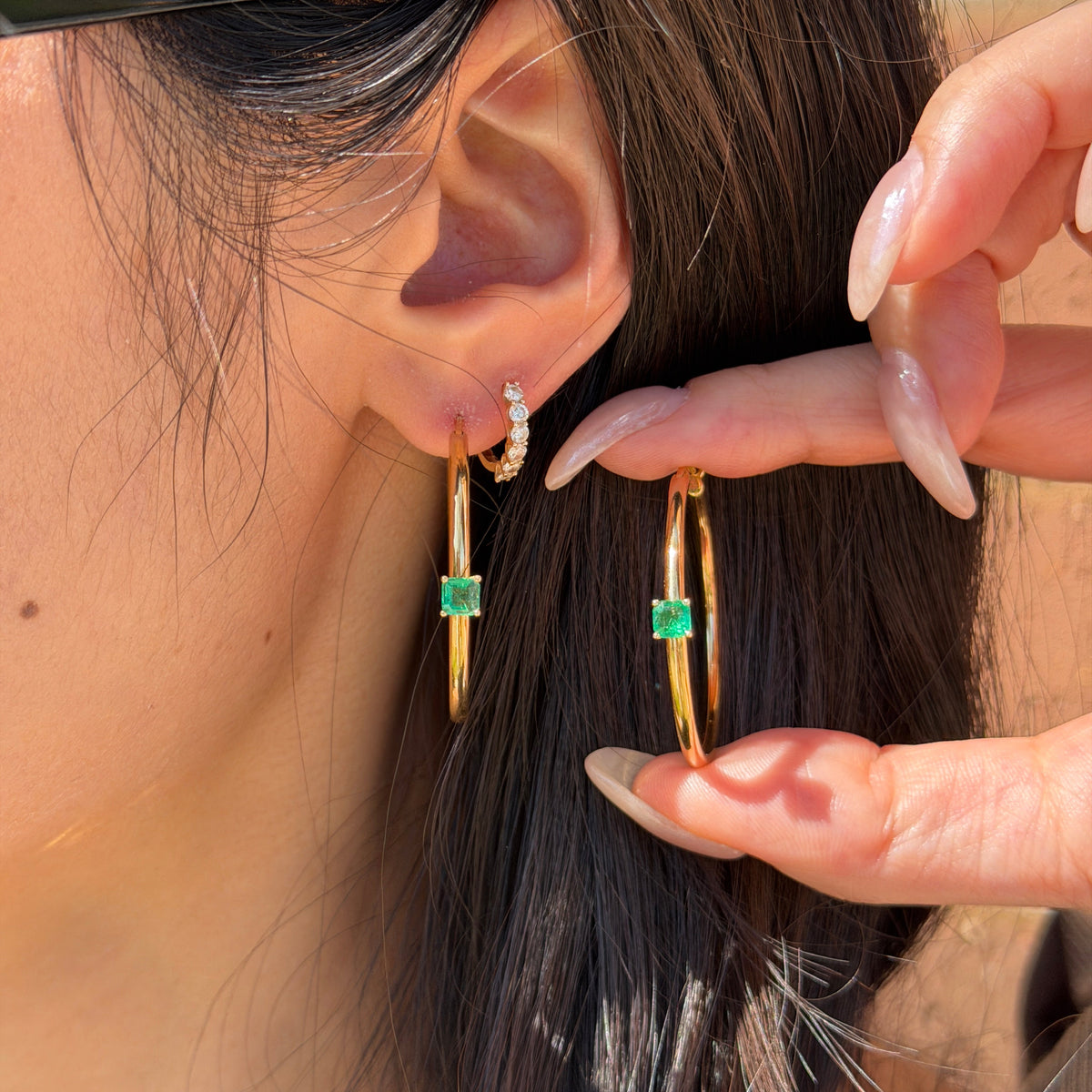 Model wearing 18K yellow gold large hoop earrings featuring a center-set vibrant green cushion-cut emerald station.