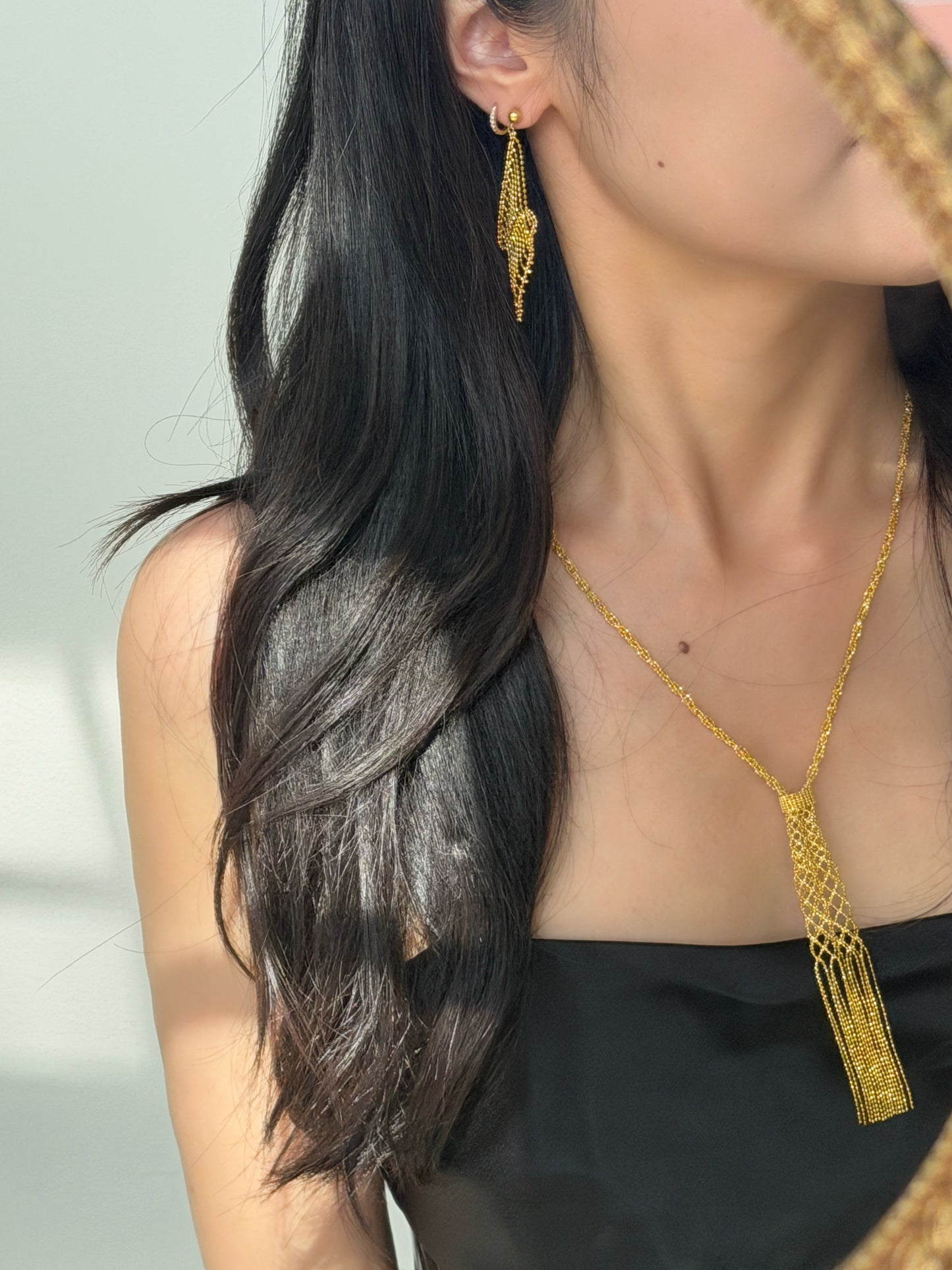 A close-up of a person with long, dark wavy hair wearing a black strapless top, modeling a delicate gold chain necklace with a woven mesh tassel pendant and a matching gold drop earring.