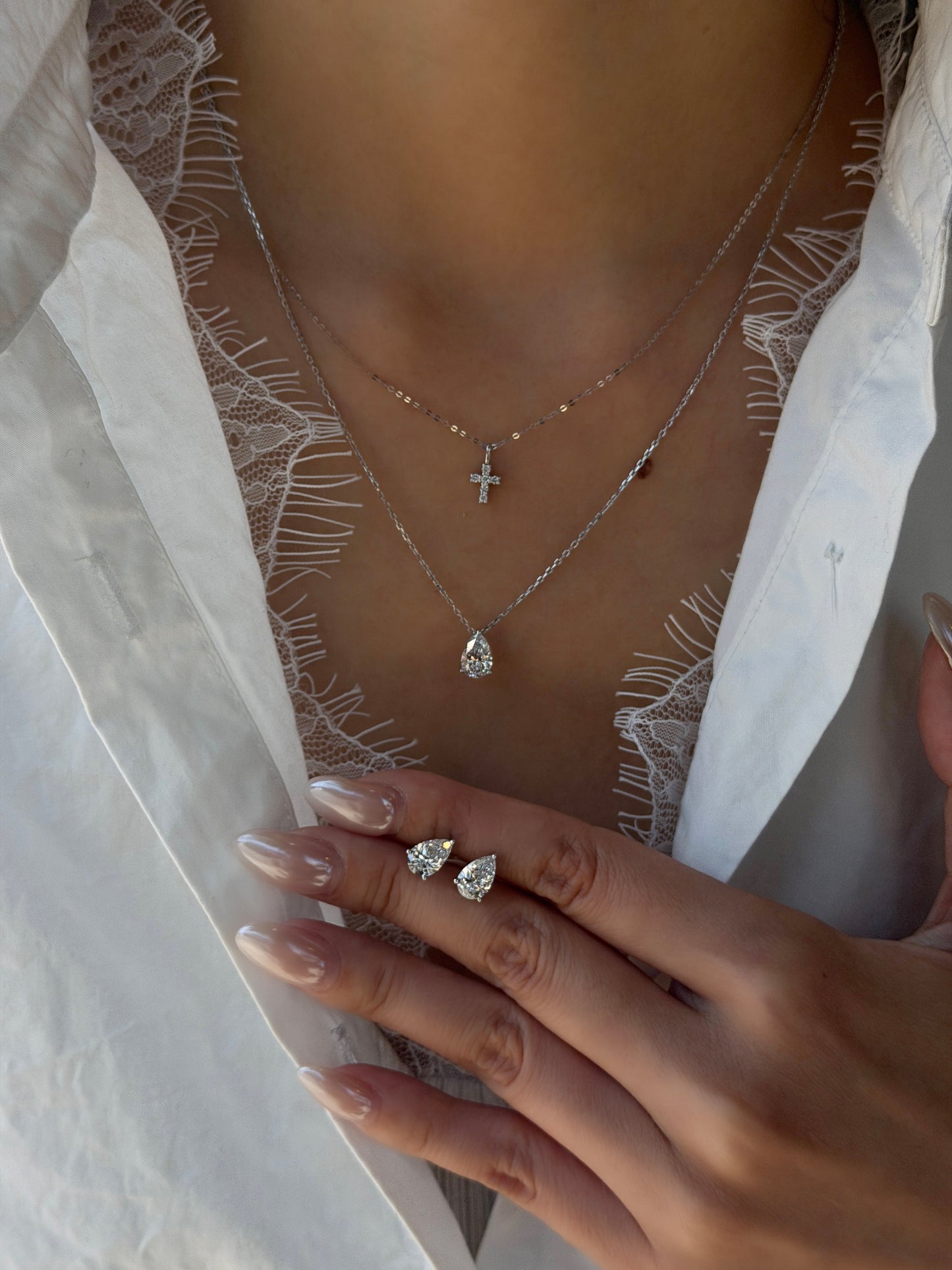 Close-up of a model wearing a 1-carat pear-cut lab-grown diamond pendant necklace on a silver chain, layered with a small diamond cross necklace and matching pear-shaped diamond stud earrings.