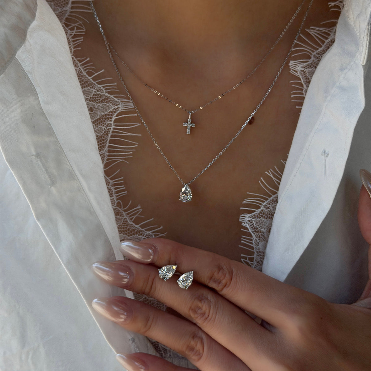 Close-up of a model wearing a 1-carat pear-cut lab-grown diamond pendant necklace on a silver chain, layered with a small diamond cross necklace and matching pear-shaped diamond stud earrings.