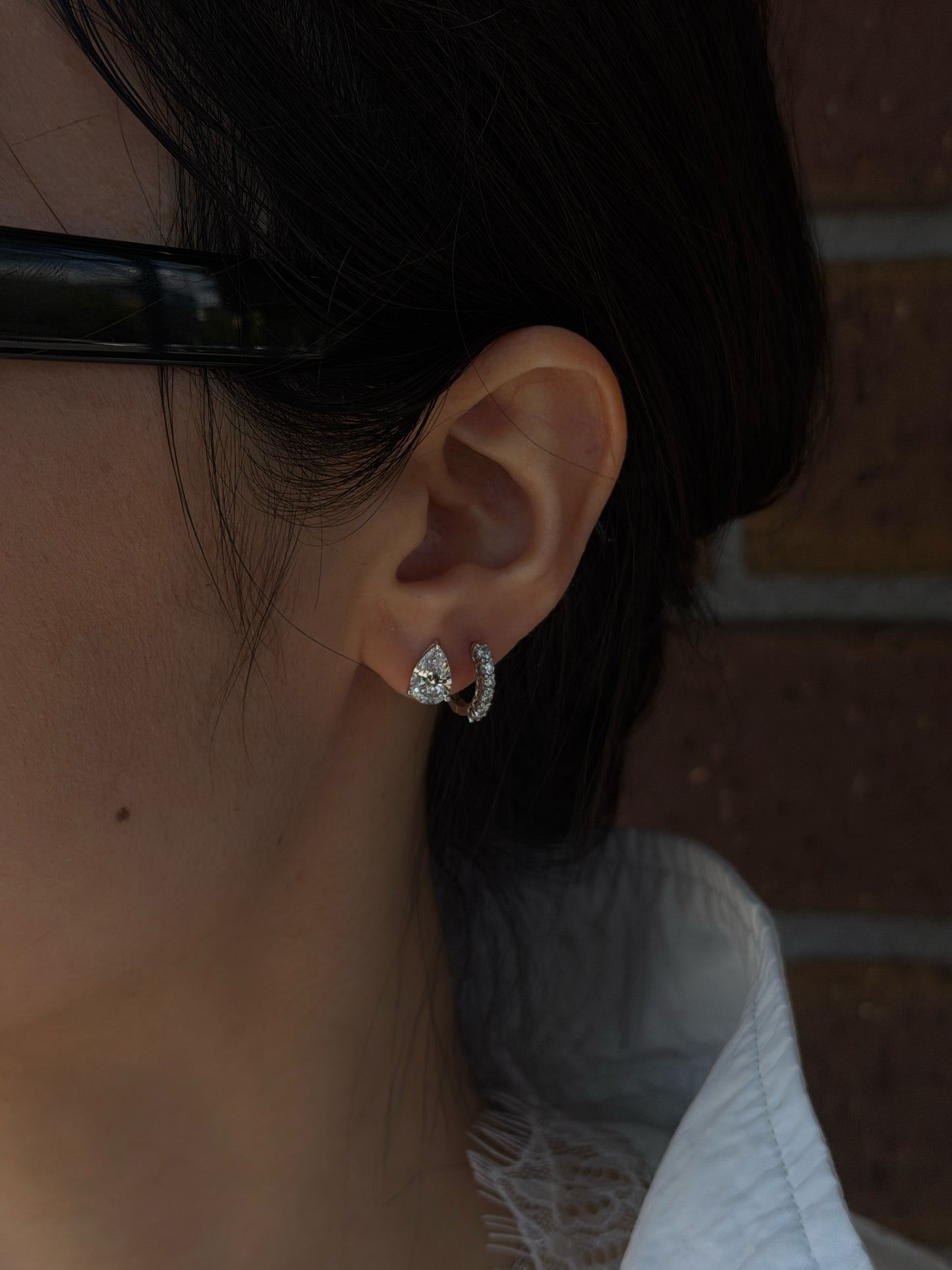 A close-up profile shot of a person's ear showcasing two diamond earrings. The primary lobe piercing features a prominent pear-shaped diamond stud, while the adjacent second piercing holds a smaller diamond-encrusted huggie hoop earring. The person has dark hair gently pulled back, with a softly focused brick wall in the background