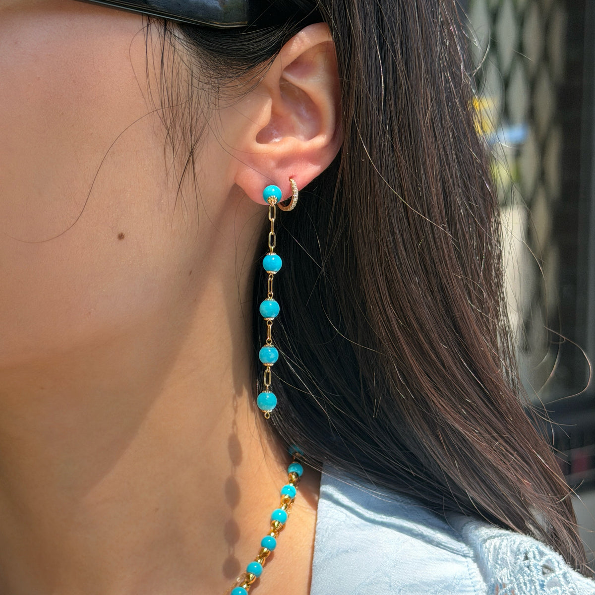 A close-up of a woman's side profile, focusing on her ear and neck. She is wearing a long dangle earring featuring bright blue Persian turquoise beads connected by gold chain links, alongside a small diamond hoop in a second piercing. The matching Persian turquoise and gold bead necklace is visible around her neck. She is wearing a light blue shirt.
