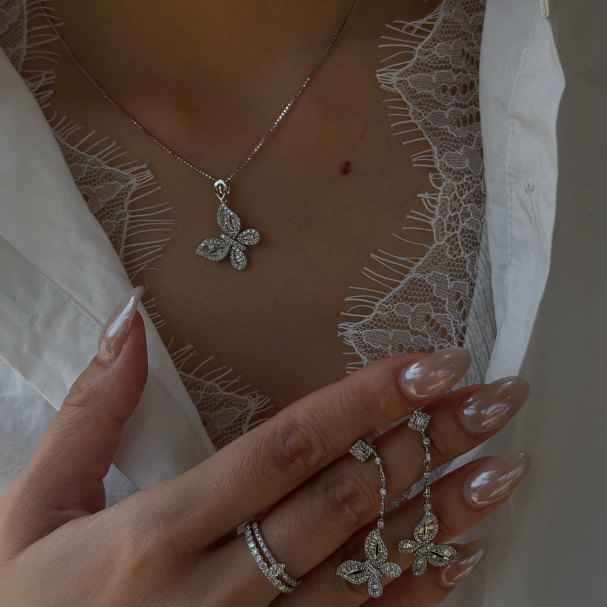 Close-up of a person wearing a white lace top under an open white shirt, modeling an 18K white gold diamond butterfly necklace. Their hand is resting near their chest, holding a matching pair of diamond butterfly dangle earrings, featuring long pearlescent nails and a diamond double-band ring.