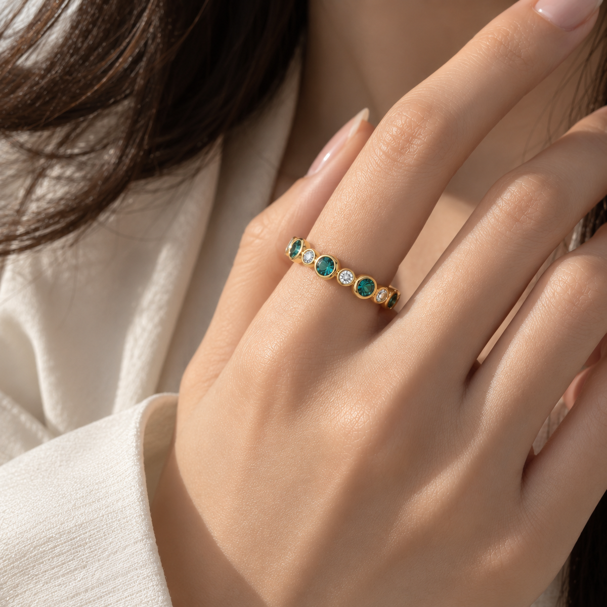 A close-up of a woman's hand resting on light beige fabric, showcasing a gold band ring featuring alternating small round tourmaline stones and diamonds.