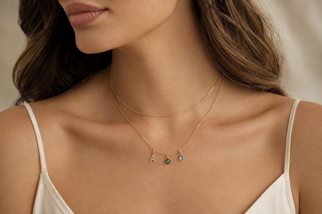 Adult model wearing layered gold necklaces with a diamond initial, emerald and aquamarine birthstone charms.