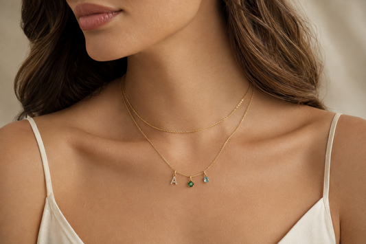 Adult model wearing layered gold necklaces with a diamond initial, emerald and aquamarine birthstone charms.
