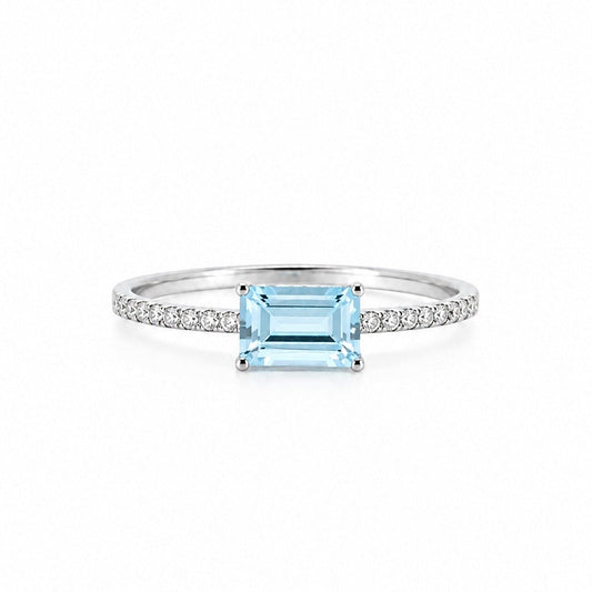 An elegant 18K white gold fine jewelry ring featuring a horizontally set emerald-cut light blue aquamarine center stone on a delicate pavé diamond band, isolated against a crisp white background.