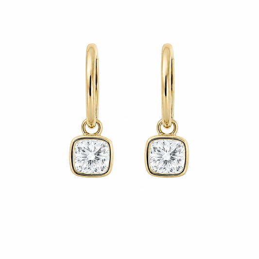 A pair of modern 18K yellow gold fine jewelry huggie hoop earrings, each featuring a dangling 1-carat cushion-cut lab-grown diamond center stone secured in a smooth bezel setting, isolated against a solid white background.