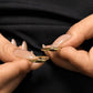 A close-up photograph showing two slender, polished 18k gold rings with a subtle wave design, held between the thumb and index finger of two separate hands with long, beige-lacquered fingernails. The hands and rings are positioned against a black textured fabric background.