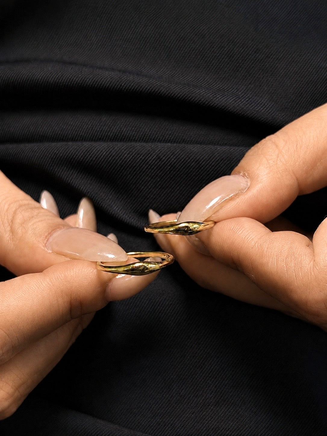 A close-up photograph showing two slender, polished 18k gold rings with a subtle wave design, held between the thumb and index finger of two separate hands with long, beige-lacquered fingernails. The hands and rings are positioned against a black textured fabric background.