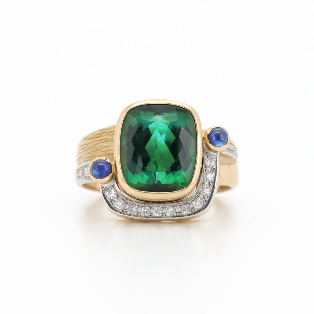 A luxury 18K two-tone gold statement ring featuring an 8.52-carat cushion-cut green tourmaline bezel-set above a pavé diamond crescent, flanked by two cabochon blue sapphires against a white background.