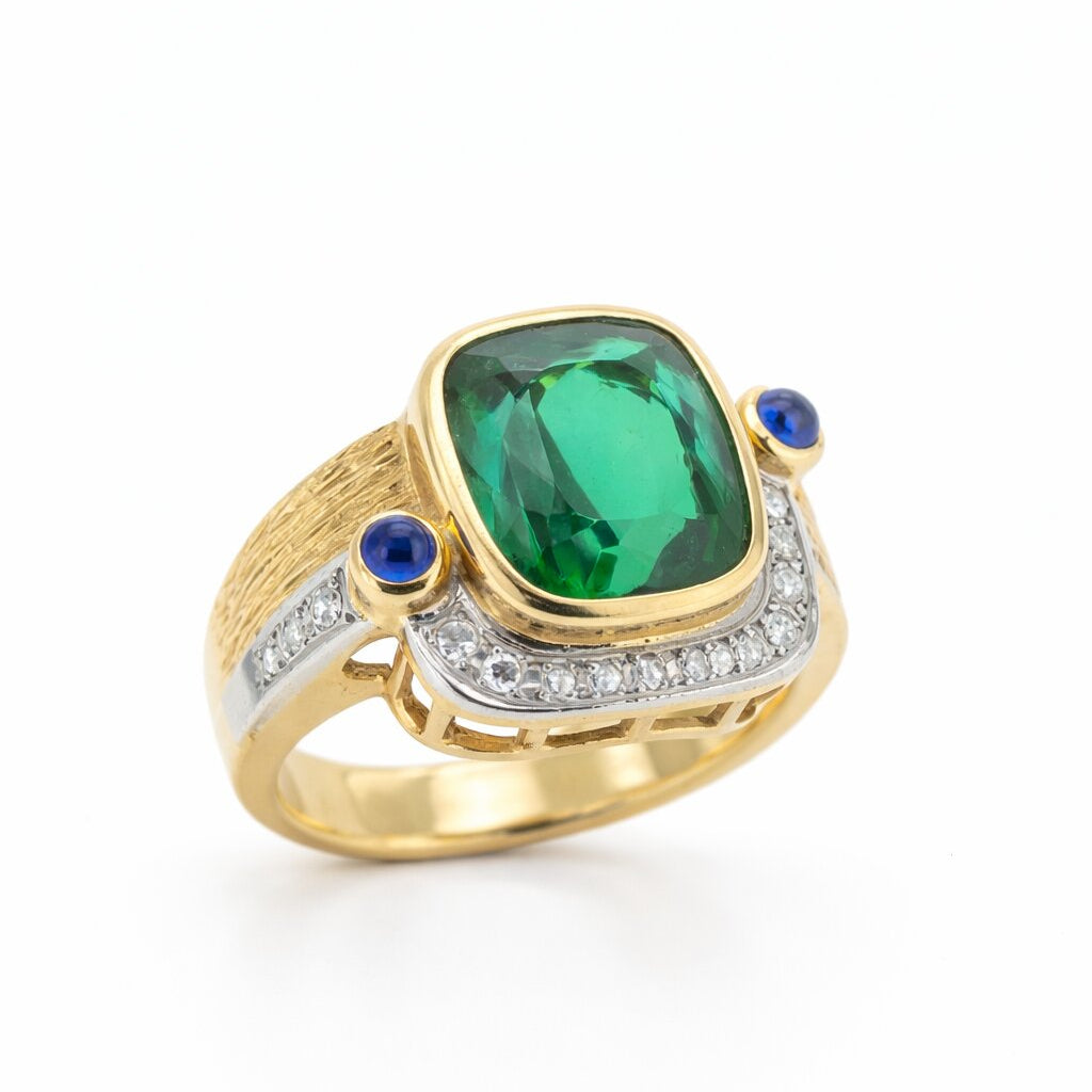 Perspective view of a high jewelry 18K gold ring displaying a large 8.52ct green tourmaline gemstone, side cabochon sapphires, and an artisanal hand-textured wire-brushed finish on the gold band.