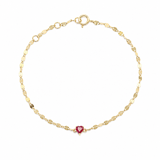 A minimalist 18K yellow gold fine jewelry bracelet featuring a small, vibrant heart-cut natural red ruby centered on a shimmering, textured Margarita chain against a solid white background.