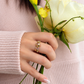 A woman wearing The Fairy Garden 18k gold kinetic ring stacked on one finger, featuring peridots and pink sapphires, holding a yellow flower.