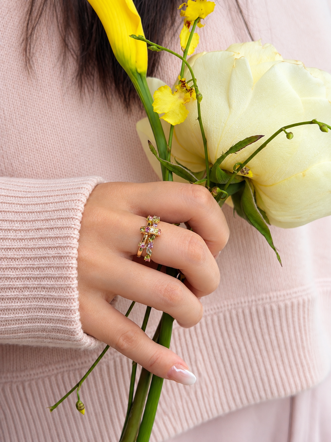 A woman wearing The Fairy Garden 18k gold kinetic ring stacked on one finger, featuring peridots and pink sapphires, holding a yellow flower.