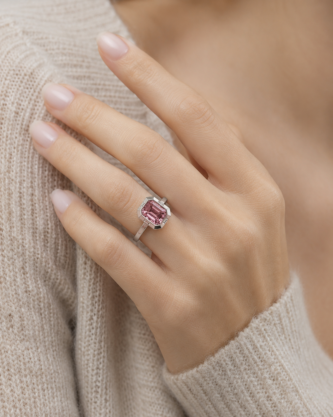 Close-up model shot of a 18k white gold ring featuring an emerald-cut natural pink tourmaline, worn on the hand against a soft beige knit sweater with no face shown.