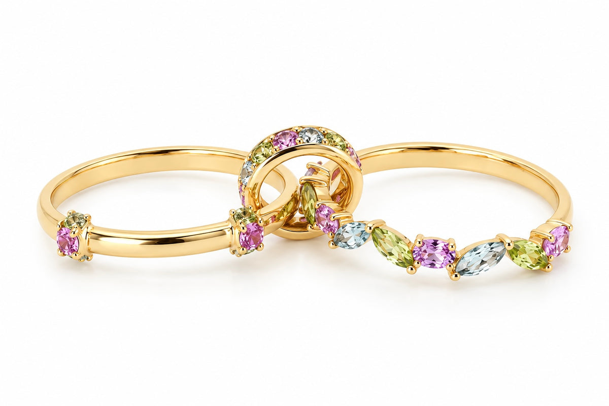 The Fairy Garden custom kinetic ring laid open against a white background. It features two 18k yellow gold bands connected by a movable center loop. The ring is beautifully set with a whimsical, vine-like pattern of green peridots, pink sapphires, and light blue aquamarines in round and marquise cuts.