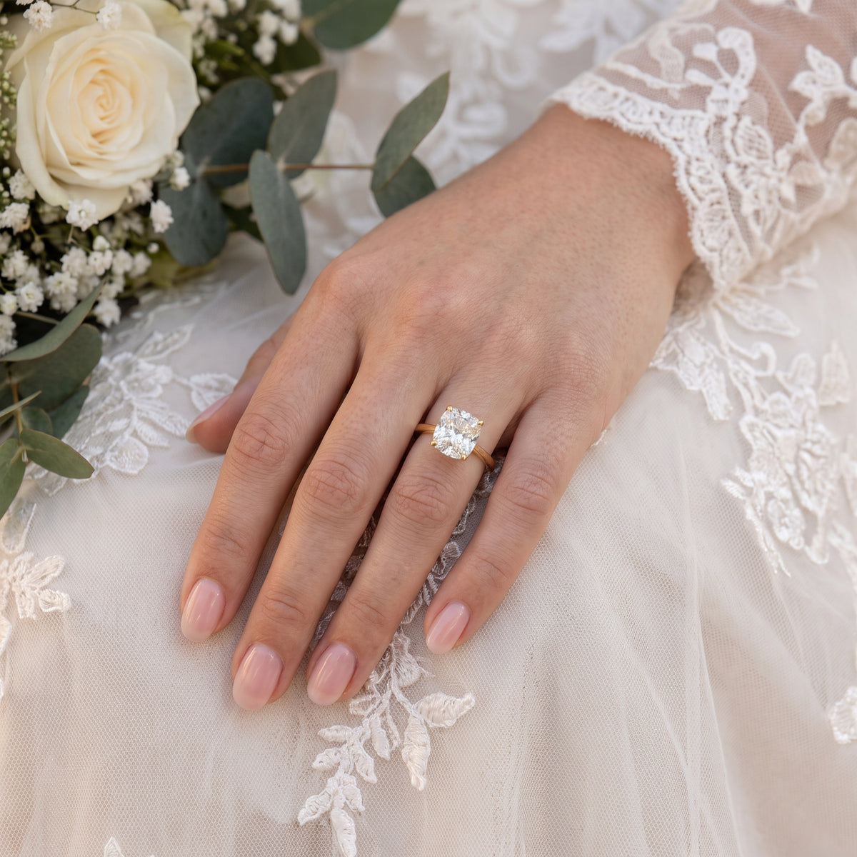 A close-up of a woman's left hand resting on a white lace wedding dress, showcasing a large cushion-cut natural diamond engagement ring on a gold band. 