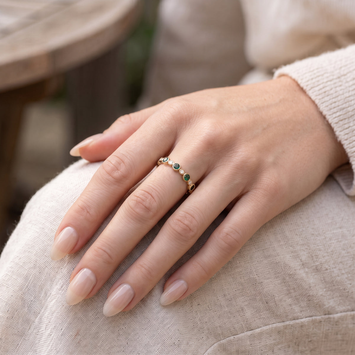 A close-up of a woman's hand resting on light beige fabric, showcasing a gold band ring featuring alternating small round tourmaline stones and diamonds. 