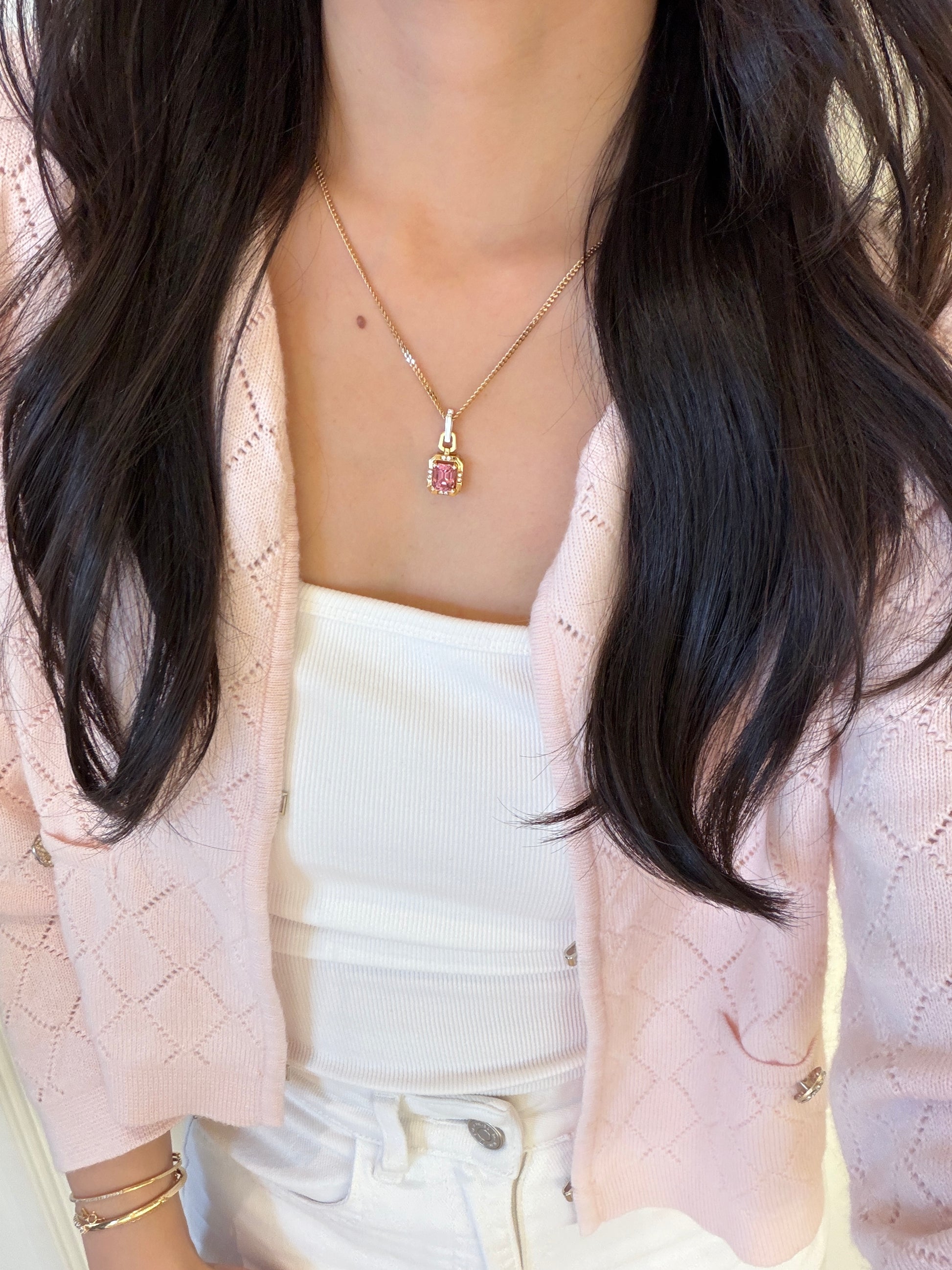 A woman wearing an 18k yellow gold necklace with a rectangular natural pink tourmaline pendant, styled with a white top and a light pink knit cardigan.