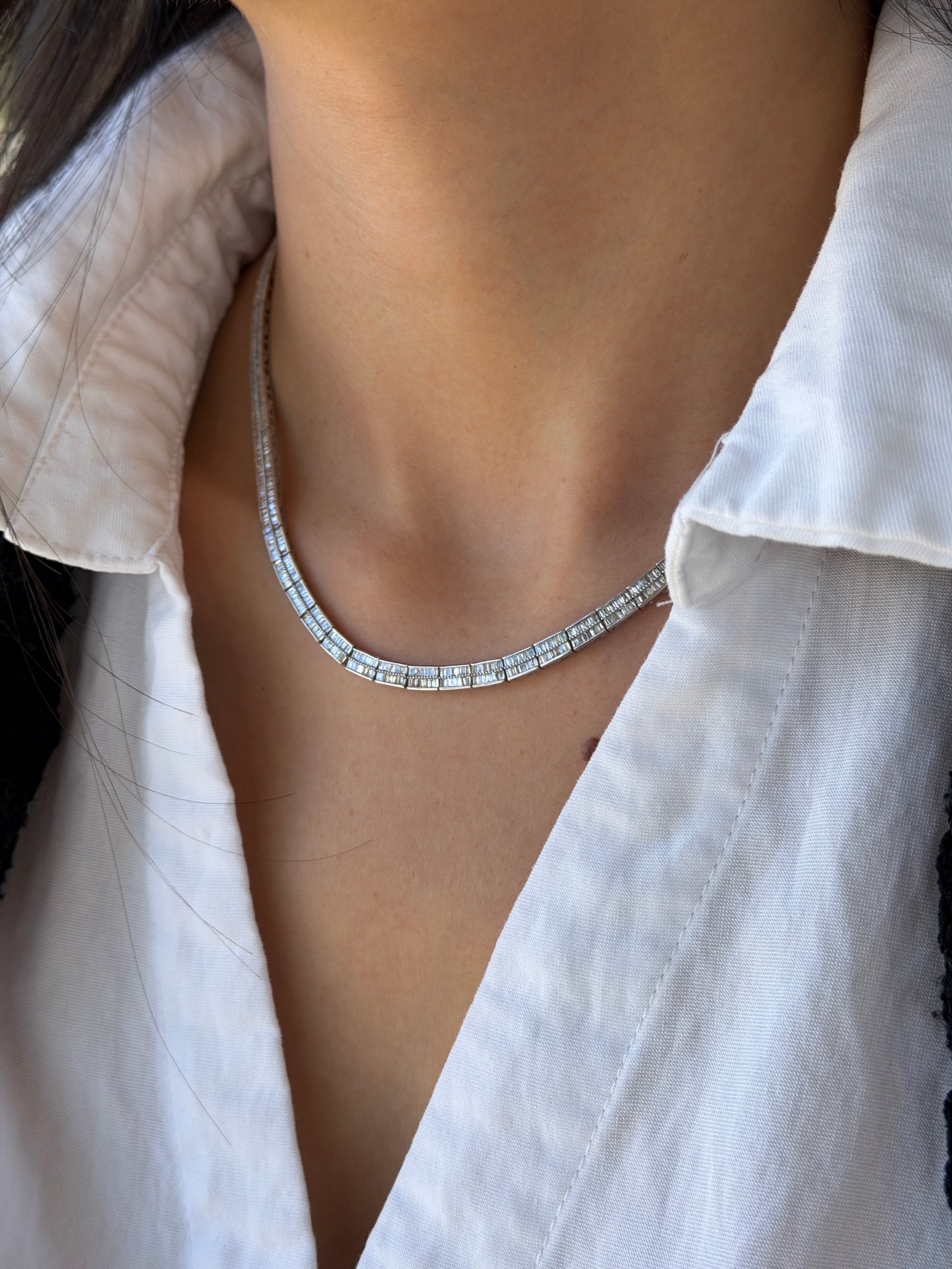 Close-up in IMG_0725.jpg of a woman wearing a sparkling baguette-cut diamond tennis necklace with an open white shirt.