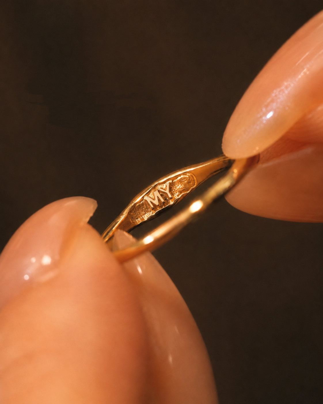 Macro photography of fingers holding an 18K gold ring, revealing the custom secret initial MY engraved on the inner band.