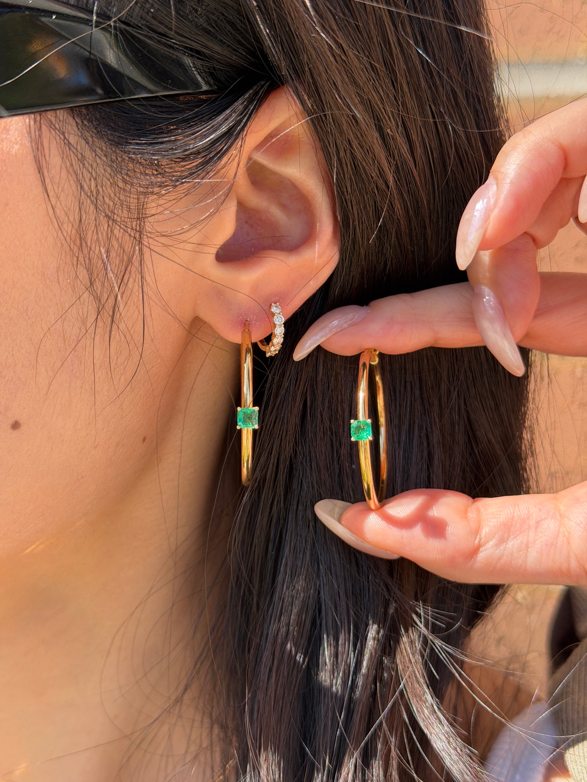 Model wearing 18K yellow gold large hoop earrings featuring a center-set vibrant green cushion-cut emerald station.