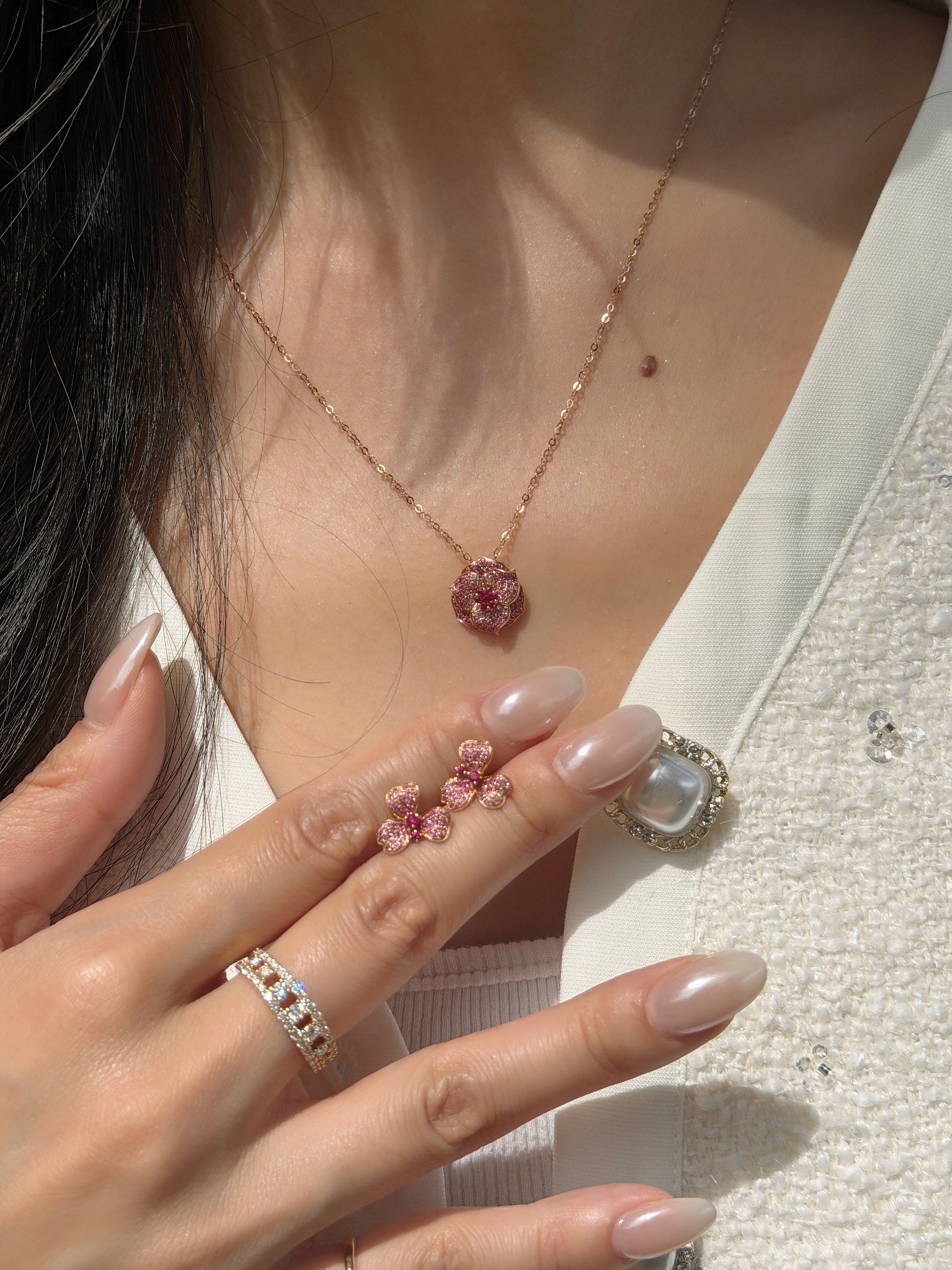 A close-up of a woman wearing an 18K natural ruby floral pendant necklace on a delicate gold chain, while holding a matching pair of ruby floral stud earrings.