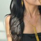 A close-up of a person with long, dark wavy hair wearing a black strapless top, modeling a delicate gold chain necklace with a woven mesh tassel pendant and a matching gold drop earring.