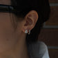 A close-up profile shot of a person's ear showcasing two diamond earrings. The primary lobe piercing features a prominent pear-shaped diamond stud, while the adjacent second piercing holds a smaller diamond-encrusted huggie hoop earring. The person has dark hair gently pulled back, with a softly focused brick wall in the background