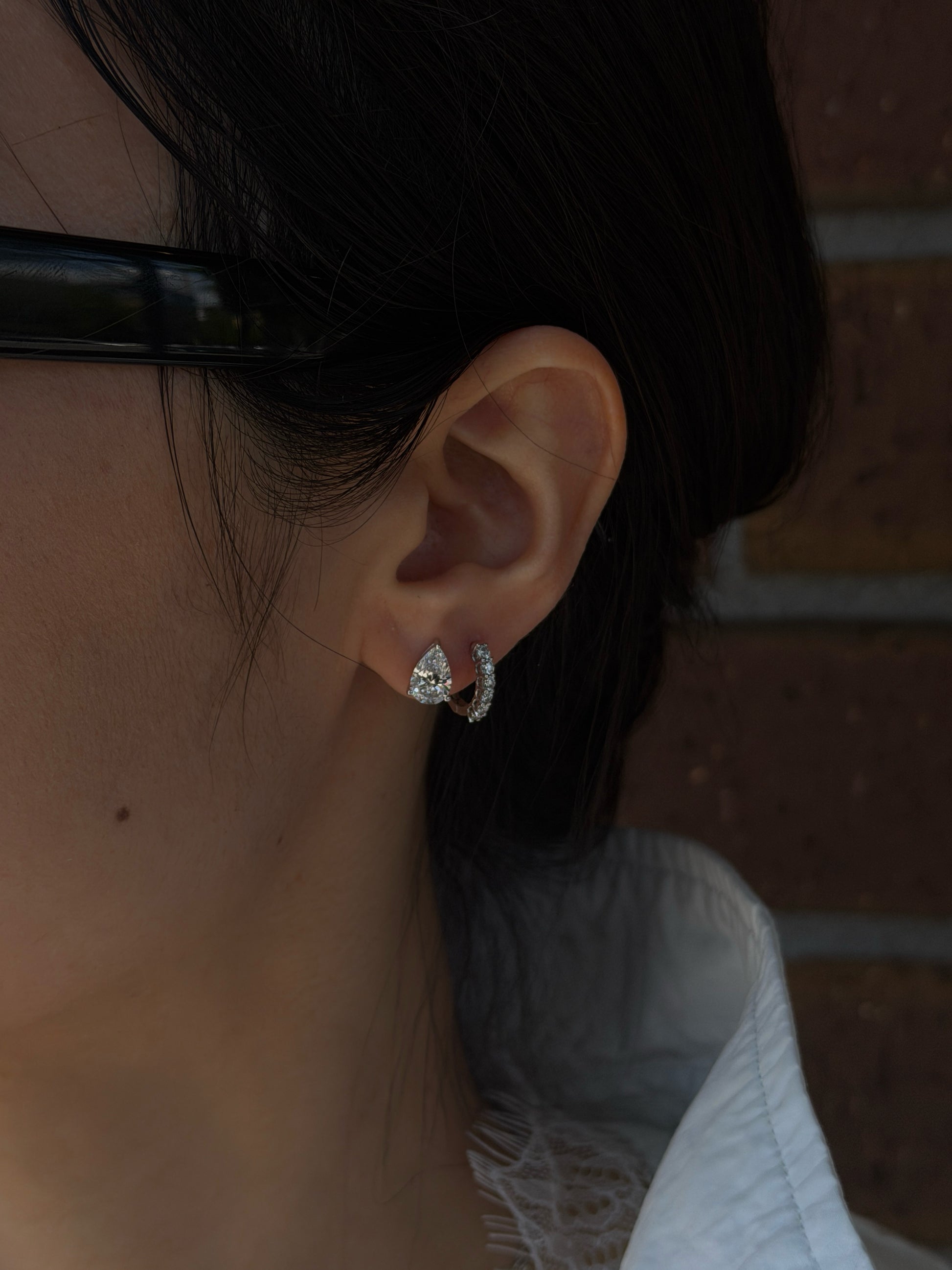 A close-up profile shot of a person's ear showcasing two diamond earrings. The primary lobe piercing features a prominent pear-shaped diamond stud, while the adjacent second piercing holds a smaller diamond-encrusted huggie hoop earring. The person has dark hair gently pulled back, with a softly focused brick wall in the background