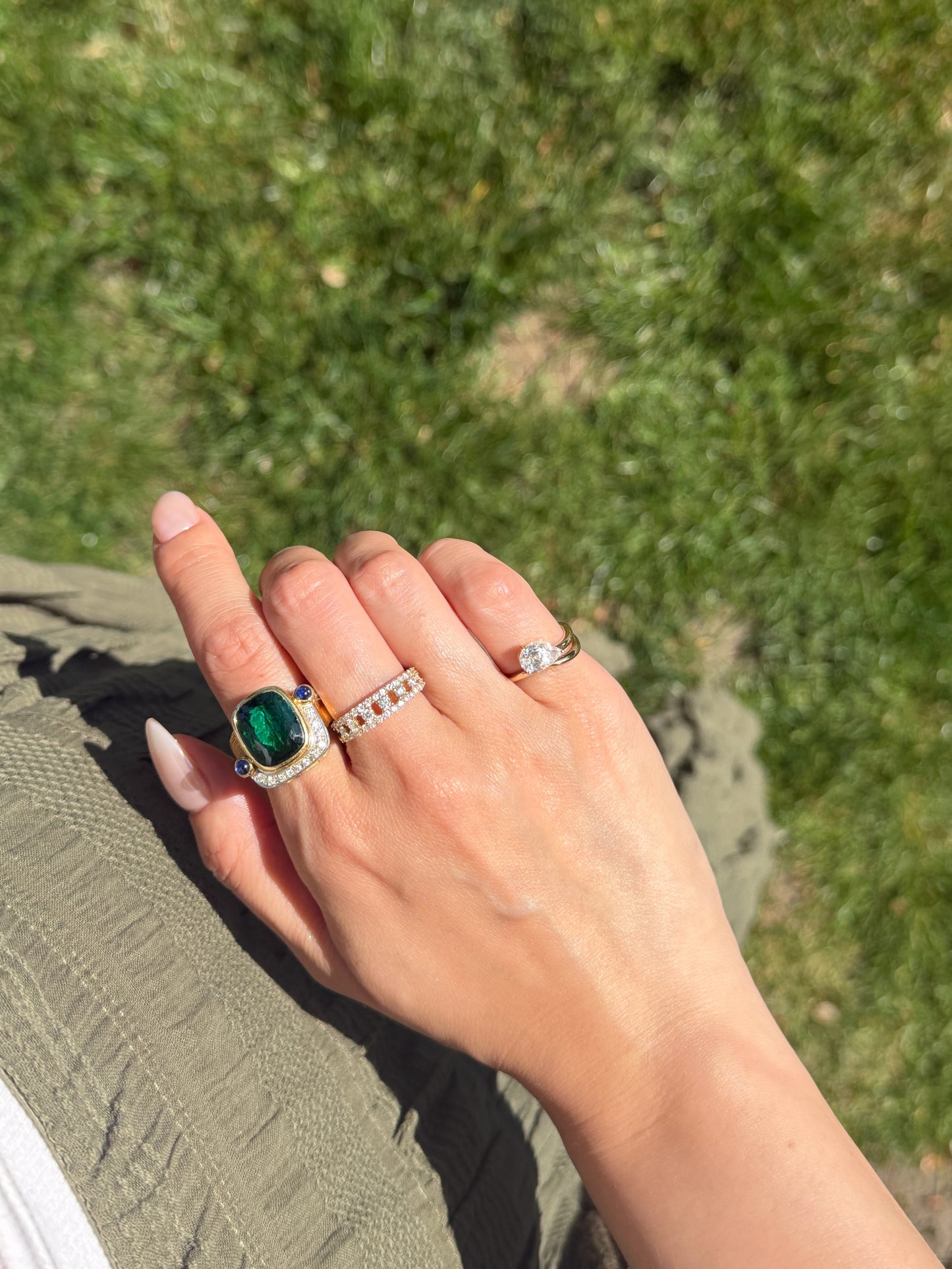 Hand wearing an 8.52 carat natural green tourmaline ring with diamond halo, blue sapphire accents, and an 18K gold setting, styled outdoors in natural sunlight.