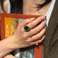 Close-up lifestyle photo of an 8.52 carat natural green tourmaline, diamond, and sapphire ring in 18K gold, worn with diamond bands and photographed against a book in warm sunlight.