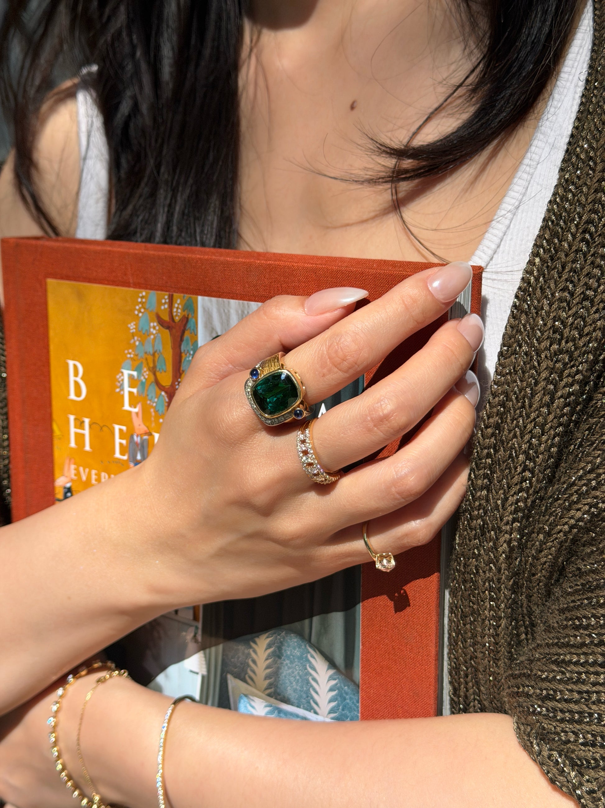 Close-up lifestyle photo of an 8.52 carat natural green tourmaline, diamond, and sapphire ring in 18K gold, worn with diamond bands and photographed against a book in warm sunlight.