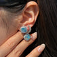 A close-up of a woman's ear and hand. Her fingers are gently resting near her earlobe, displaying a square-cut opal earring with vibrant blue and green play-of-color, surrounded by a halo of small diamonds. She is also wearing a small, diamond-encrusted hoop earring in a second piercing.