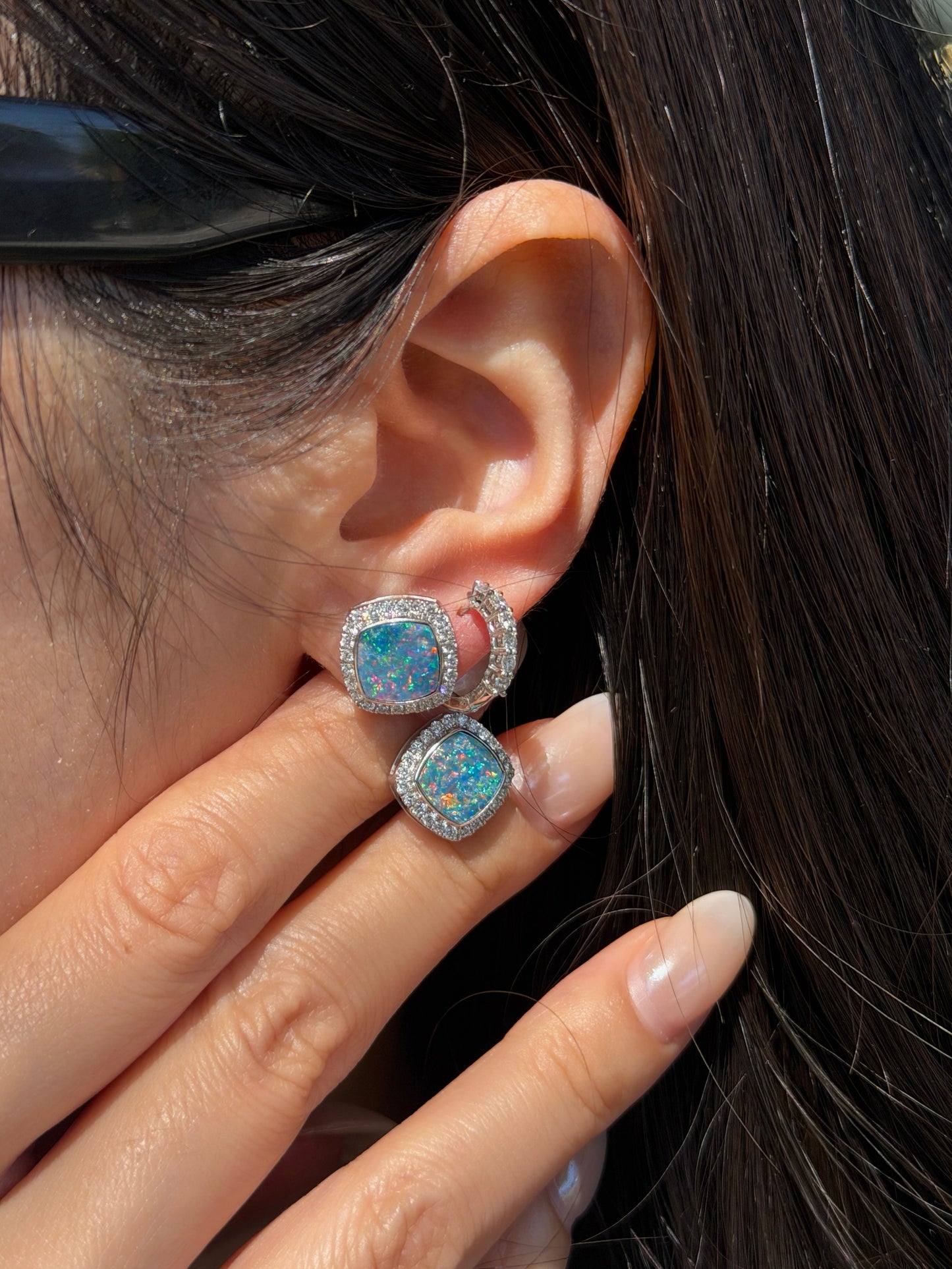 A close-up of a woman's ear and hand. Her fingers are gently resting near her earlobe, displaying a square-cut opal earring with vibrant blue and green play-of-color, surrounded by a halo of small diamonds. She is also wearing a small, diamond-encrusted hoop earring in a second piercing.