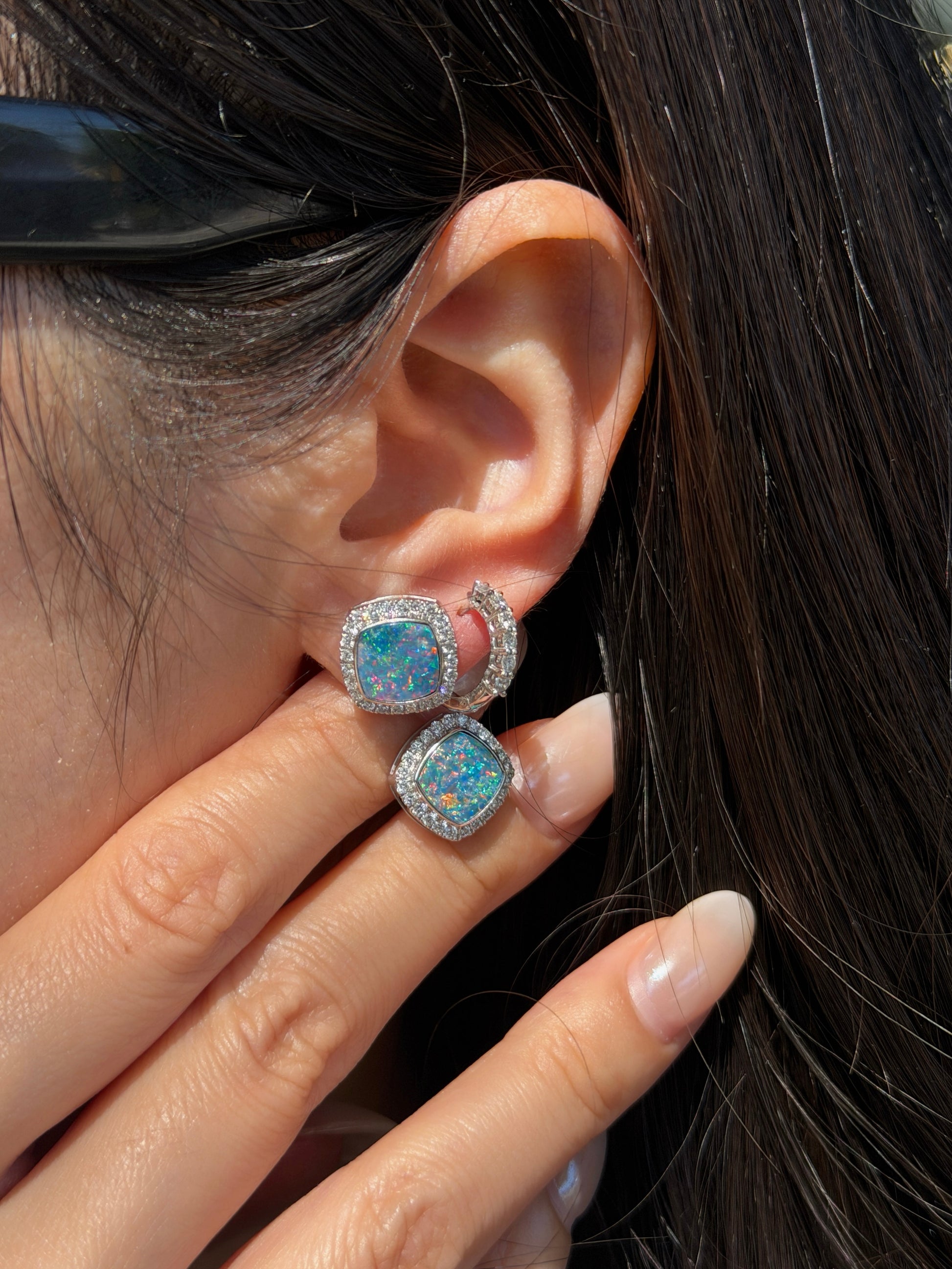 A close-up of a woman's ear and hand. Her fingers are gently resting near her earlobe, displaying a square-cut opal earring with vibrant blue and green play-of-color, surrounded by a halo of small diamonds. She is also wearing a small, diamond-encrusted hoop earring in a second piercing.