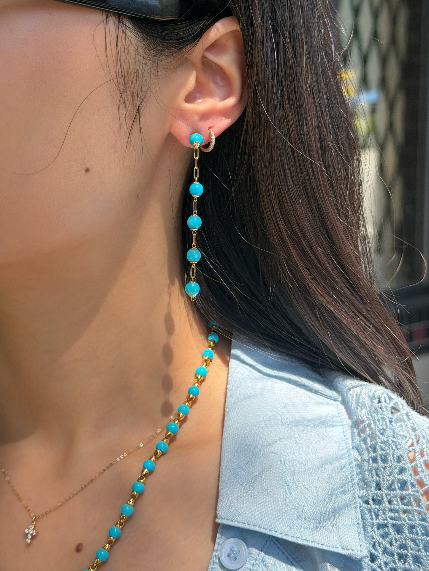 A close-up of a woman's side profile, focusing on her ear and neck. She is wearing a long dangle earring featuring bright blue Persian turquoise beads connected by gold chain links, alongside a small diamond hoop in a second piercing. The matching Persian turquoise and gold bead necklace is visible around her neck. She is wearing a light blue shirt.