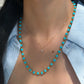 A close-up of a woman's neck and chest showcasing layered necklaces. She is wearing a long necklace made of alternating gold and bright blue Persian turquoise beads, paired with a shorter, delicate gold chain featuring a small diamond cross pendant. She is wearing a light blue button-down shirt slightly open over a white top.
