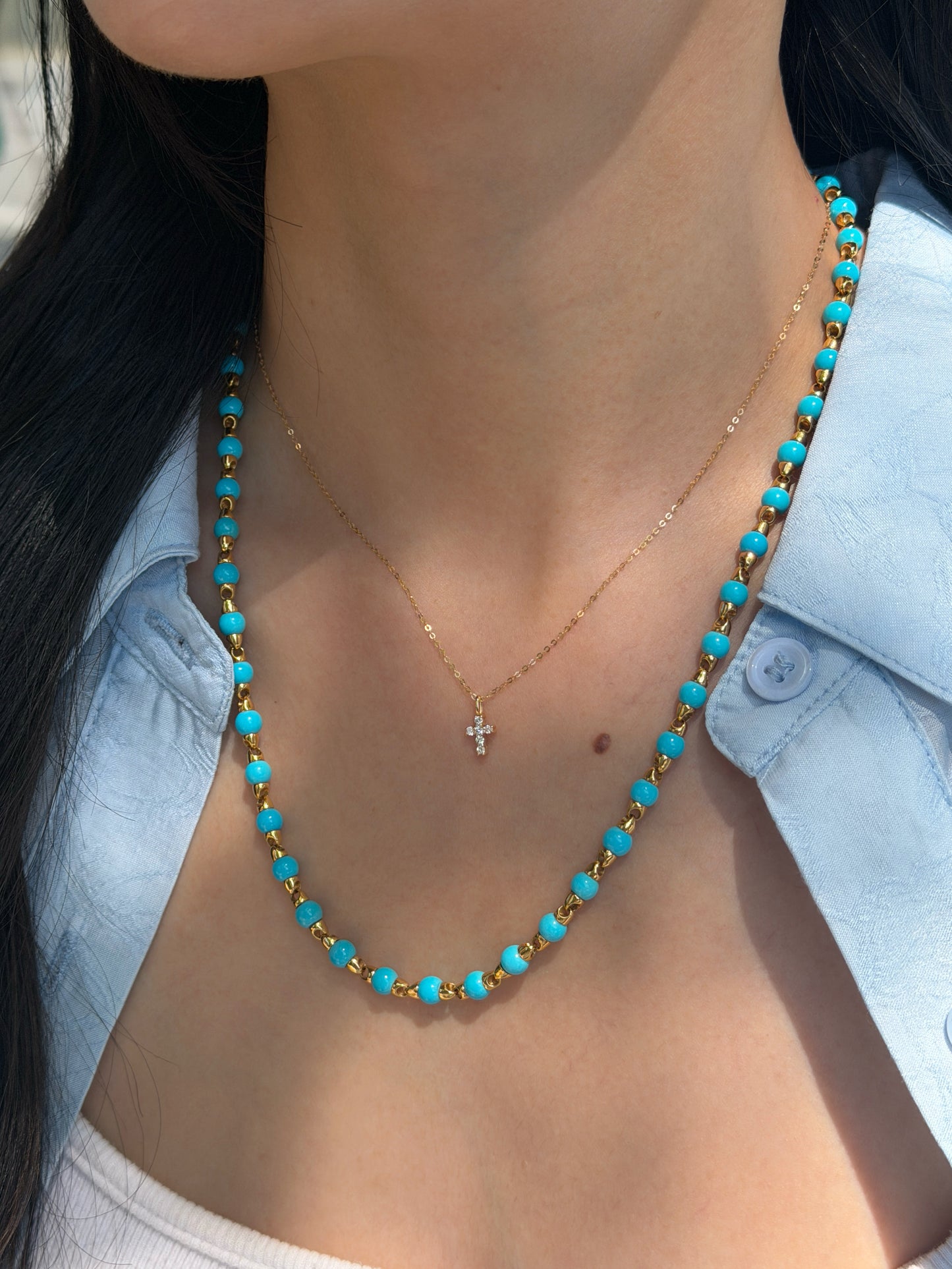 A close-up of a woman's neck and chest showcasing layered necklaces. She is wearing a long necklace made of alternating gold and bright blue Persian turquoise beads, paired with a shorter, delicate gold chain featuring a small diamond cross pendant. She is wearing a light blue button-down shirt slightly open over a white top.
