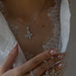 Close-up of a person wearing a white lace top under an open white shirt, modeling an 18K white gold diamond butterfly necklace. Their hand is resting near their chest, holding a matching pair of diamond butterfly dangle earrings, featuring long pearlescent nails and a diamond double-band ring.