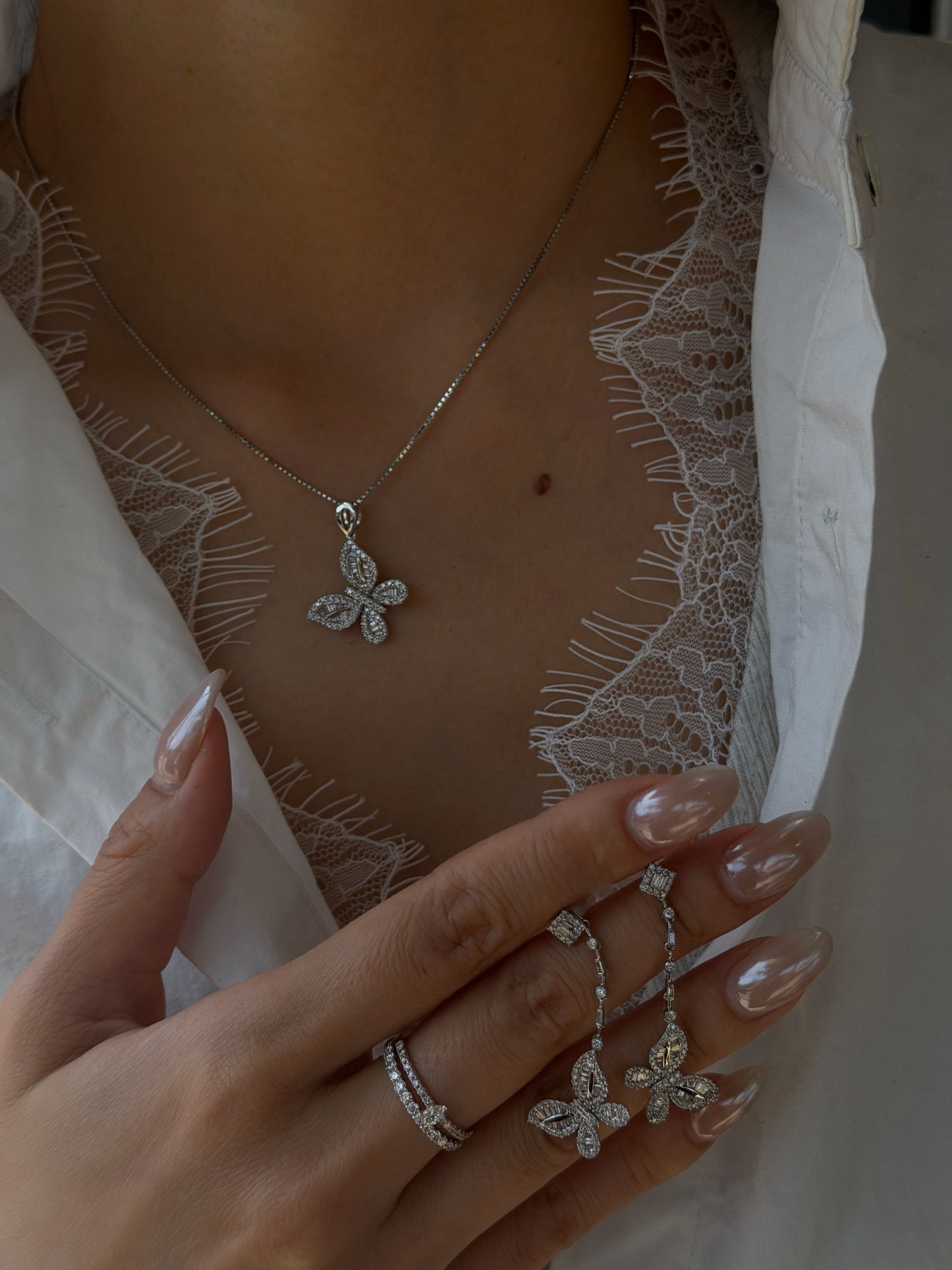 Close-up of a person wearing a white lace top under an open white shirt, modeling an 18K white gold diamond butterfly necklace. Their hand is resting near their chest, holding a matching pair of diamond butterfly dangle earrings, featuring long pearlescent nails and a diamond double-band ring.