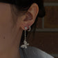 Close-up lifestyle image of a person's ear modeling an 18K white gold diamond butterfly drop earring. The earring hangs from the primary earlobe piercing, showing the articulated drop chain and diamond butterfly motif, and is paired with a small diamond huggie hoop in the second piercing. The background is a softly blurred dark brick wall.
