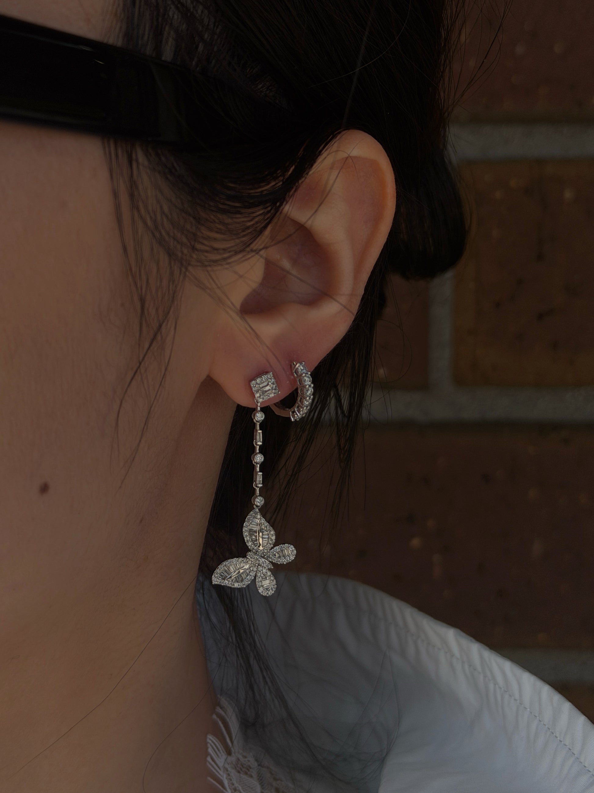 Close-up lifestyle image of a person's ear modeling an 18K white gold diamond butterfly drop earring. The earring hangs from the primary earlobe piercing, showing the articulated drop chain and diamond butterfly motif, and is paired with a small diamond huggie hoop in the second piercing. The background is a softly blurred dark brick wall.
