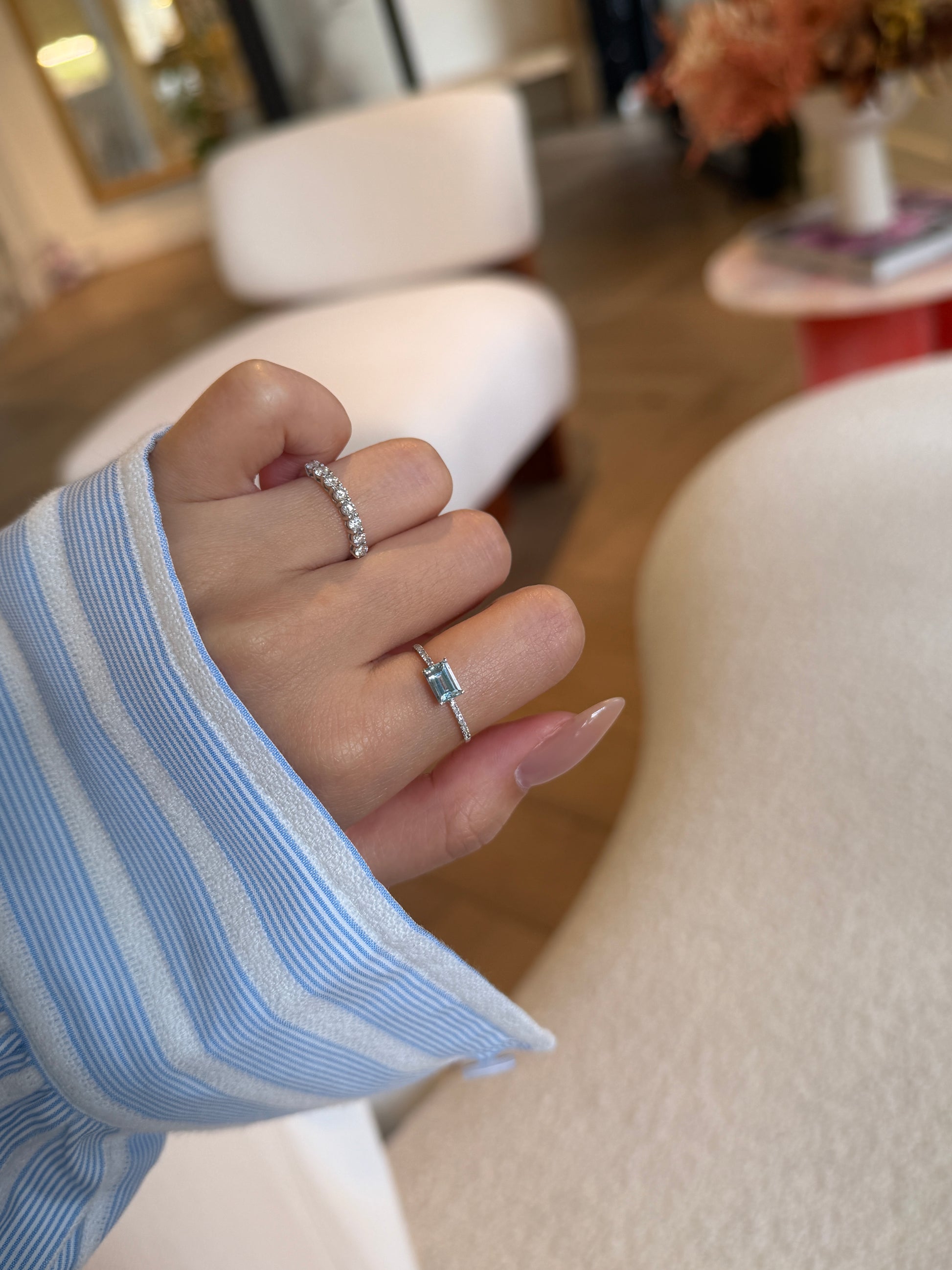 A close-up of a hand modeling two rings in a bright, modern boutique setting. The index finger features a classic diamond band, while the ring finger showcases a dainty ring with a horizontally set rectangular light blue gemstone on a thin diamond pavé band.