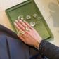 Model wearing two-tone gold and silver daisy rings and a matching chain bracelet from the Artisanal Petals collection, with matching earrings, rings, and a necklace displayed on a green velvet tray.