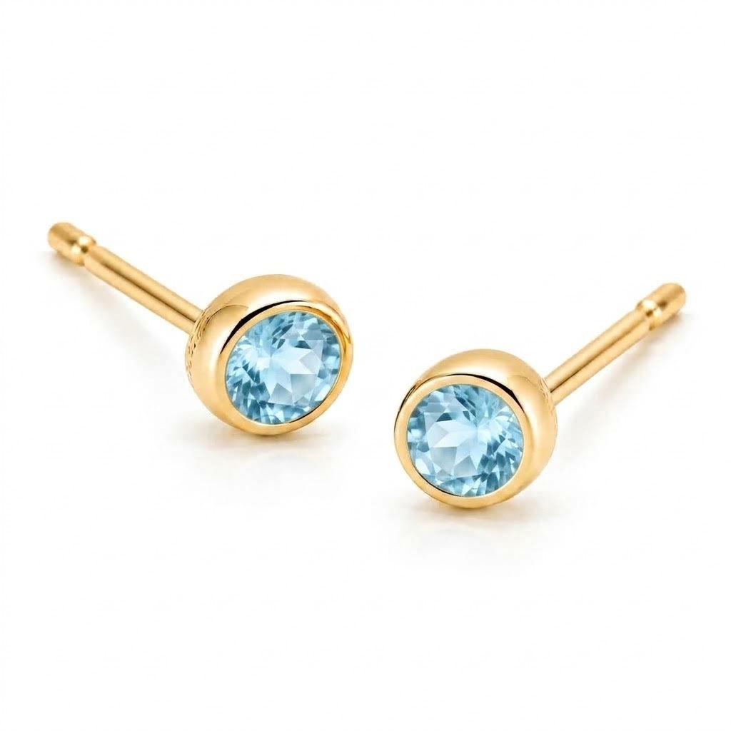 Pair of 18K yellow gold stud earrings featuring bezel-set round light blue topaz gemstones.