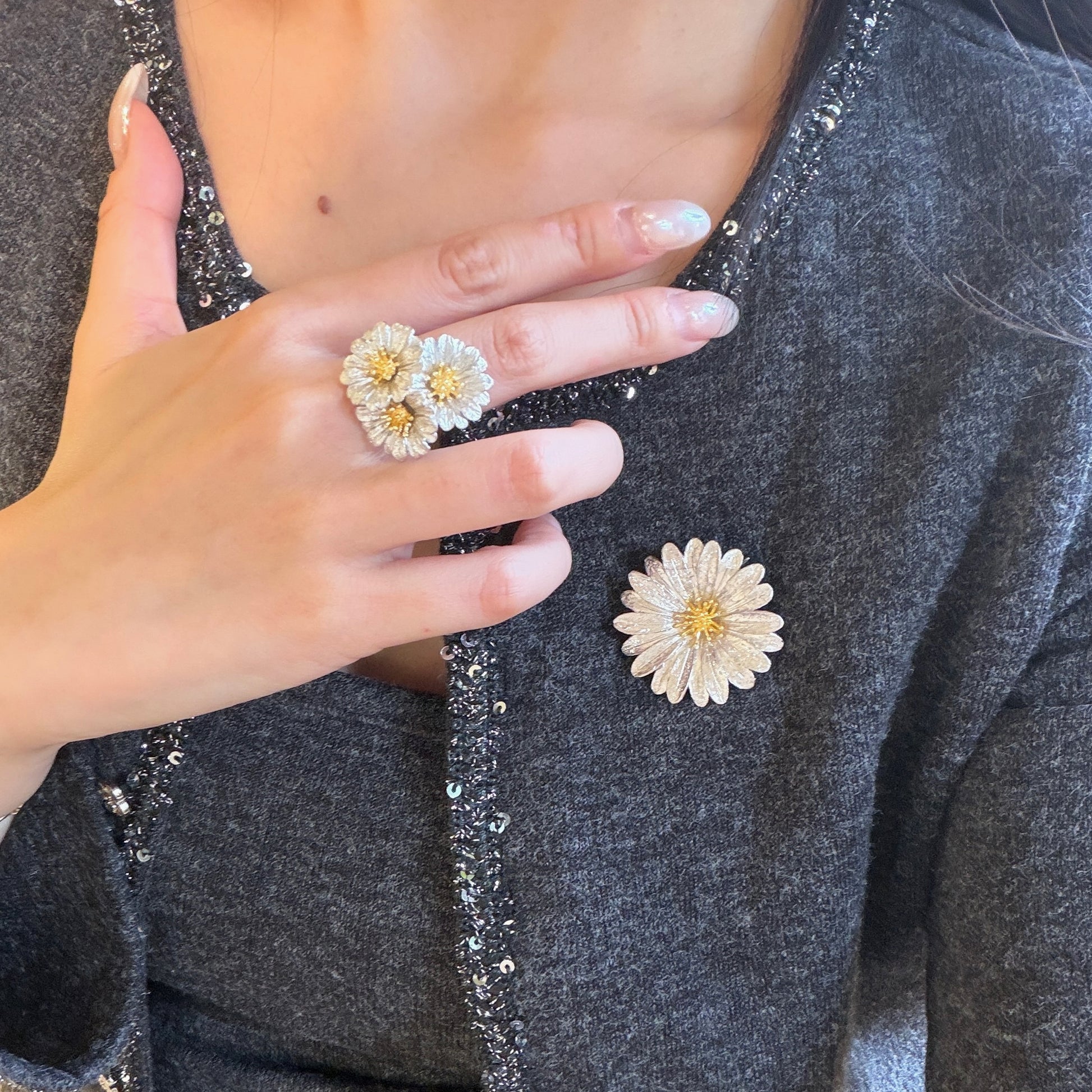 Italian silver floral jewelry featuring a textured daisy brooch with a gold-tone center and a matching three-flower statement ring, styled on a dark tailored jacket.